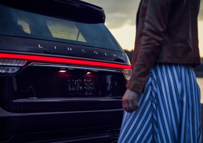 A person is shown near the rear of a 2024 Lincoln Aviator® SUV as the Lincoln Embrace illuminates the rear lights | Gates Ford Lincoln in Richmond KY
