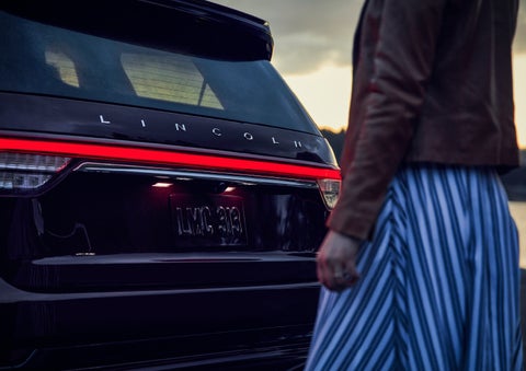 A person is shown near the rear of a 2024 Lincoln Aviator® SUV as the Lincoln Embrace illuminates the rear lights | Gates Ford Lincoln in Richmond KY